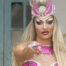 En Drag Queen i pink krystalbesat kjole og krone peger mod beskueren med et scepter