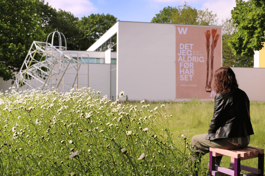 En kvinde med skulderlangt, brunt hår i sort læderjakke sidder på en farverig skammel i en park med mange hvide blomster. Hun sidder med ryggen til kameraet. Foran hende er en hvid bygning med en stor plakat for en udstilling med titlen "Det jeg aldrig før hat set".