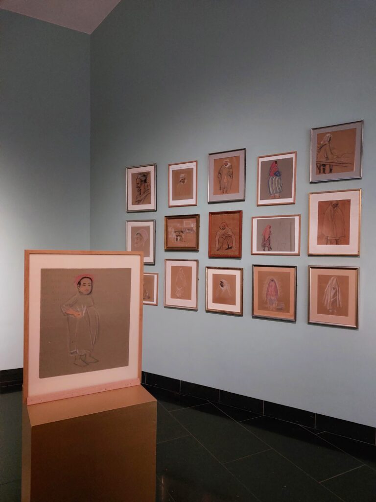 Installationsfoto fra udstillingen "I lyset af Nordafrika" på Willumsens Museum. En samling skitser i forskellige rammer er ophængt på en lyseblå væg. I forgrunden er en skitse af et barn på en piedestal.